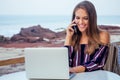 Young sexy woman in a stylish striped jumpsuit using laptop and telephone freelancing on the tropical paradise beach Royalty Free Stock Photo