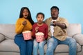 Young shocked black parents and their daughter watching movie on TV, eating popcorn, sitting on couch on blue background Royalty Free Stock Photo