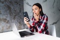 Young shocked woman sitting indoors laptop computer chatting by mobile phone Royalty Free Stock Photo