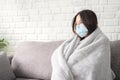 Young sick asian woman covered with blanket sits upset on sofa at home, wearing medical protective mask, looks away Royalty Free Stock Photo