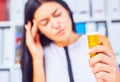 Young sick tired sad woman sitting in workplace holding bottle with pills. Female feeling bad at work.