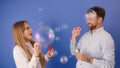 Young silly caucasian couple blowing bubbles and having fun. Isolated on blue background