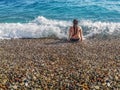 A young slender woman in a bikini sits among the ocean waves on a pebble beach Royalty Free Stock Photo