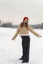 Young slender woman in black jeans and beige sheepskin coat with red knitted hat walks in snow outdoors. Winter walk