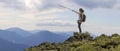 Young slim blond tourist girl with backpack points with stick at foggy mountain range panorama standing on rocky top on bright blu