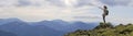 Young slim blond tourist girl with backpack points with stick at foggy mountain range panorama standing on rocky top on bright