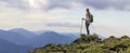 Young slim blond tourist girl with backpack and stick standing on rocky top against bright blue morning sky on foggy mountain rang