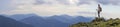 Young slim blond tourist girl with backpack and stick standing on rocky top against bright blue morning sky on foggy mountain