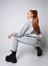 Young slim red-haired woman in sportswear hoodie, pants and black massive shoes sits squatted back to camera
