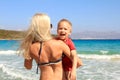 Young slim woman holding her son on the beach by the clear blue sea. Little boy in UV filter clothes is smiling, mother and her