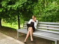 Young slim woman talking on the phone and looking into the laptop while sitting on a bench in the park. Cute adult girl in a Royalty Free Stock Photo