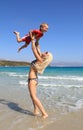 Young slim woman throws up and catches her little son on the beach by the clear blue sea. Mother and her cute little baby-boy are