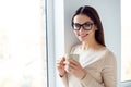 Young smart successful smiling woman in glasses having break, holding cup and drink coffe while standing near window