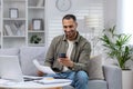 Young smiling African-American man sitting on sofa at home, holding phone and documents, working on laptop Royalty Free Stock Photo