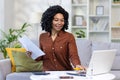 Young smiling African American woman working from home with documents and bills, sitting on sofa at home and using Royalty Free Stock Photo