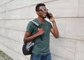 Young smiling african man student calling on smartphone wearing backpack, sunglasses walking in city over gray wall Royalty Free Stock Photo