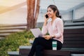 Young smiling Asian girl using a mobile phone and laptop on her lap in university campus building