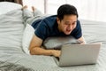 Young smiling Asian man in casual clothing and lying on bed with laptop computer. Male freelancer working from home on Royalty Free Stock Photo