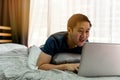 Young smiling Asian man in casual clothing and lying on bed with laptop computer. Male freelancer working from home on Royalty Free Stock Photo