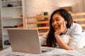 Young smiling Asian woman in casual clothing and lying on bed with laptop computer. Female freelancer working from home Royalty Free Stock Photo