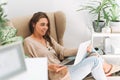 Young smiling attractive woman in cozy beige cardigan working at laptop sitting in chair at the home with green house plants Royalty Free Stock Photo