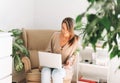 Young smiling attractive woman in cozy beige cardigan working at laptop sitting in chair at the home with green house plants Royalty Free Stock Photo