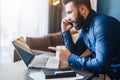 Young bearded businessman is sitting at table in front of computer,talking on cell phone,showing finger on laptop screen Royalty Free Stock Photo