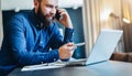 Smiling bearded businessman sitting at table in front of computer, talking on cell phone, showing pen on laptop screen Royalty Free Stock Photo