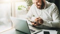 Young smiling bearded businessman in white shirt sitting at desk in front of laptop, making notes in notebook. Royalty Free Stock Photo