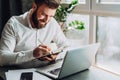 Young smiling bearded businessman in white shirt sitting at desk in front of laptop, making notes in notebook. Royalty Free Stock Photo