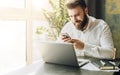 Young smiling bearded businessman sitting at table in front of computer, using smartphone. Man checking email, chatting. Royalty Free Stock Photo