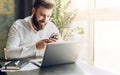 Young smiling bearded businessman sitting at table in front of computer, using smartphone. Man checking email, chatting. Royalty Free Stock Photo