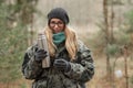 Young smiling beautiful blond woman in camouflage outfit and green scarf posing with thermos in the forest. Travel lifestyle