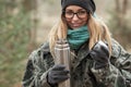 Young smiling beautiful blond woman in camouflage outfit and green scarf posing with thermos in the forest. Travel lifestyle