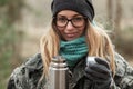 Young smiling beautiful blond woman in camouflage outfit and green scarf posing with thermos in the forest. Travel lifestyle