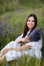 Young smiling beautiful brunette woman in white dress is sitting in green meadow with lupine flowers in spring Royalty Free Stock Photo