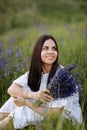 Young smiling beautiful brunette woman in white dress is sitting in green meadow with lupine flowers in spring Royalty Free Stock Photo