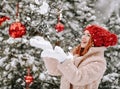 Young smiling beautiful woman in fur coat and mittens standing near decorated Christmas tree and catching snowflakes