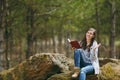 Young smiling beautiful woman sitting on stone studying reading book and spreading hands in city park or forest on green Royalty Free Stock Photo