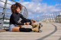 Young black girl sitting on urban bridge and puts on skates. Woman with afro hairstyle rollerblading on sunny day