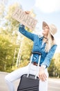 Young smiling blond woman raising looking at cardboard with inscription anywhere, sitting on black suitcase near forest. Royalty Free Stock Photo