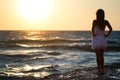 Young smiling blond woman in white dress standing on rocks and looking at sunset over wavy sea water Royalty Free Stock Photo