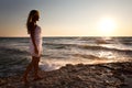 Young smiling blond woman in white dress standing on rocks and looking at sunset over wavy sea water Royalty Free Stock Photo