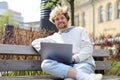 A young smiling blonde sits with a laptop on a bench in a city park Royalty Free Stock Photo