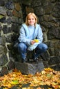 A young smiling blonde woman with yellow dandelions in her hands sits near a stone wall Royalty Free Stock Photo