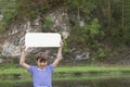 Young smiling boy standing on bank of river keeping blank white board in hands above his head, rocky cliffs on background. Summer