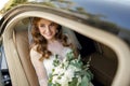 A young smiling bride sits in an expensive car and holds a white wedding bouquet of roses Royalty Free Stock Photo