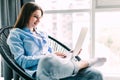 Young smiling brunette girl is sitting on modern chair near the window in light cozy room at home working on laptop in relaxing Royalty Free Stock Photo