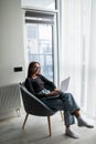 Young smiling woman sitting on modern chair near the window in light cozy room at home working on laptop in relaxing atmosphere Royalty Free Stock Photo