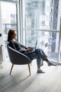 Young smiling woman is sitting on modern chair near the window in light cozy room at home. She is working on laptop in relaxing Royalty Free Stock Photo